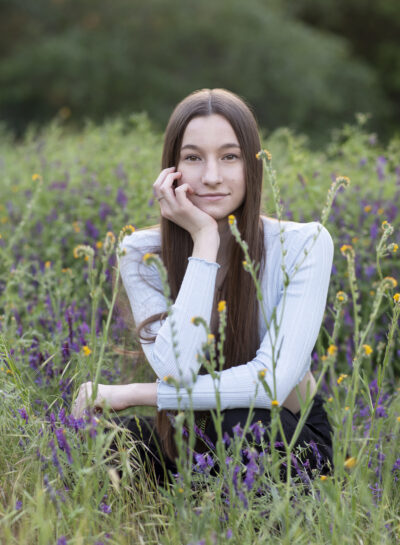 A long haired brunette teen girl crouched in a field of purple and yellow flowers.