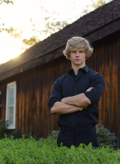 Blond high school senior boy, dressed in black, standing with folded arms, in front of a wood barn.