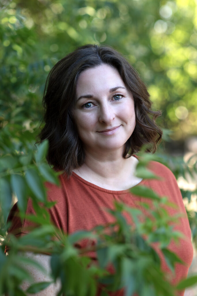 Brunette woman in an orange shirt standing among green bushes.