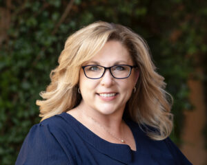 Headshot of a female realtor wearing a navy-blue blouse in front of green foliage.