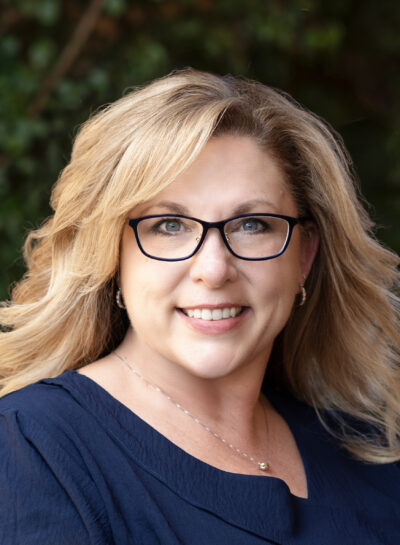 Headshot of a female realtor wearing a navy-blue blouse in front of green foliage.