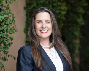 Headshot of a female realtor wearing a white blouse with a navy-blue blazer in front of green foliage.