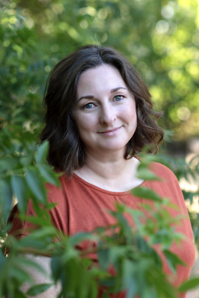 Brunette woman in an orange shirt standing among green bushes.