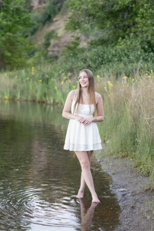 Blonde haired teen, wearing a white sundress, standing at the lakeshore.