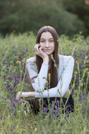 A long haired brunette teen girl crouched in a field of purple and yellow flowers.