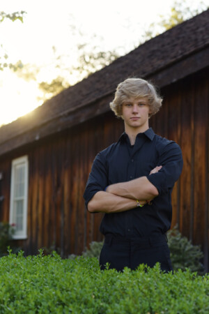 Blond high school senior boy, dressed in black, standing with folded arms, in front of a wood barn.